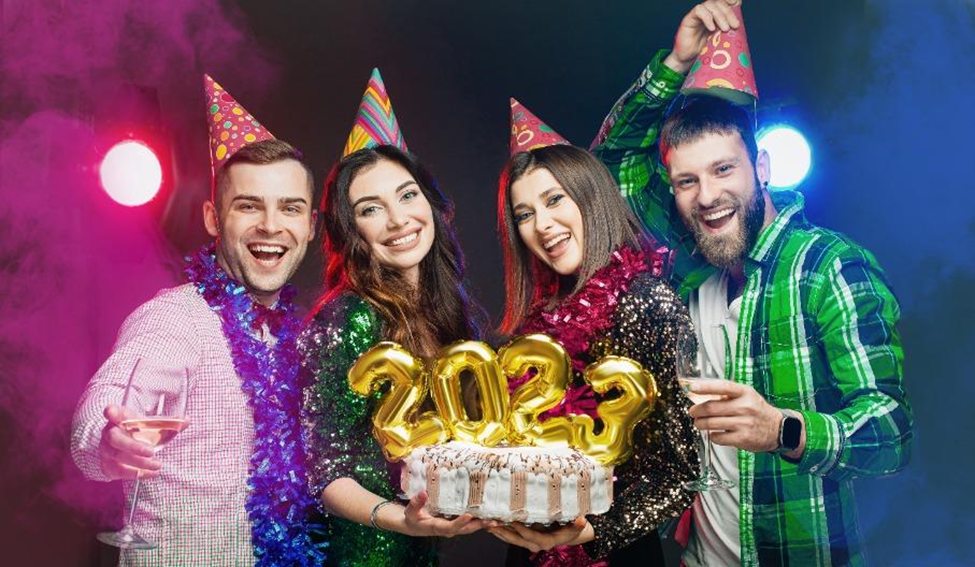 Four people wearing party hats holding a cake with 2023 number toppers on it, along with glasses of champagne.
